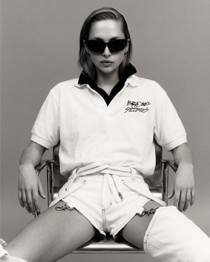 Black-and-white campaign photograph: model seated in a chrome chair wearing a Brashy Studios logo polo