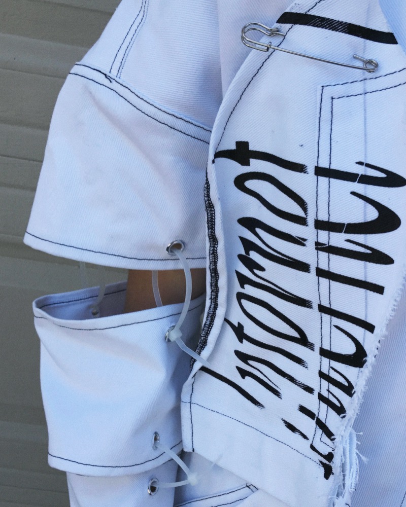 Close detail of a white garment: script print, steel safety pin, zip-tie through grommets