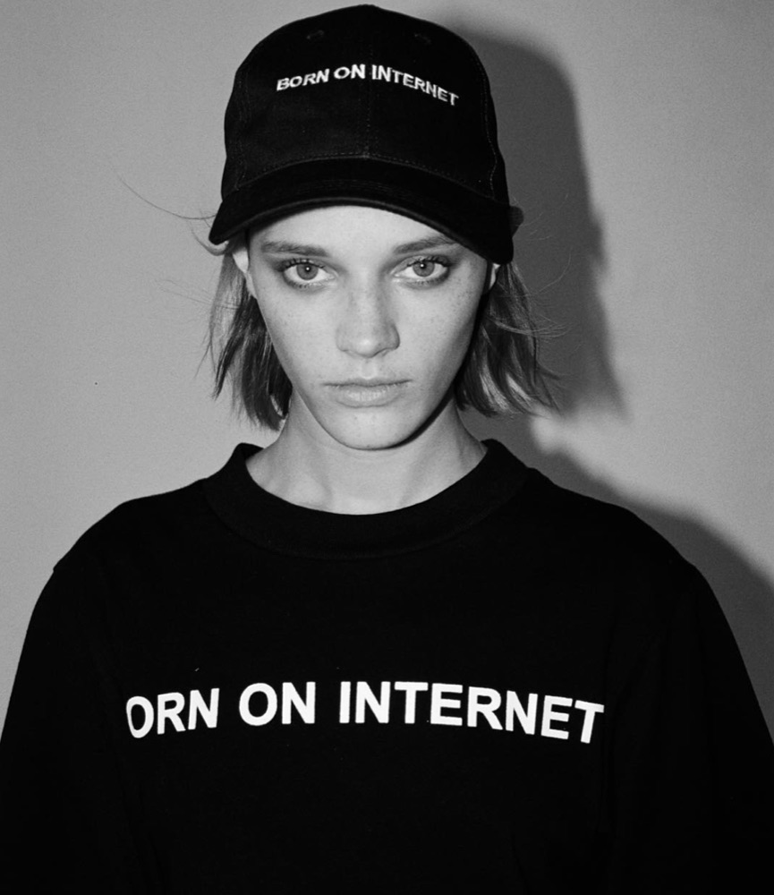 Black-and-white studio portrait: model in a black cap embroidered BORN ON INTERNET and a black crewneck reading ORN ON INTERNET, the B falling outside the frame
