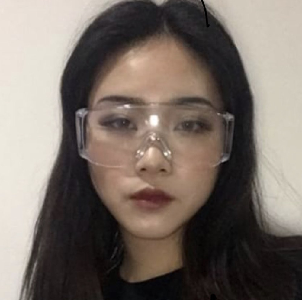 Front-camera selfie: model in clear plastic safety goggles and dark lipstick against a bare wall