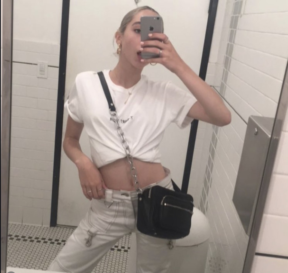 Public bathroom mirror selfie: knotted white tee, black harness strap, hanging chain, white carpenter jeans with hardware, black nylon crossbody bag