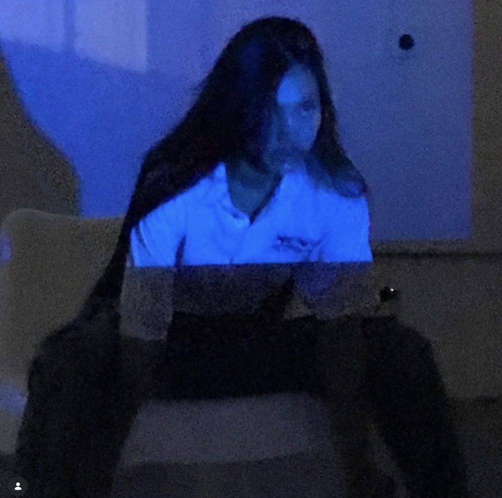 Model seated in blue projector light wearing a white logo polo; the projected image falls across her body and the wall behind, with an Instagram profile chip visible in the corner