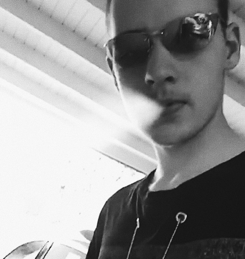 Black-and-white close selfie: young man in sunglasses and a black tee, safety pin and ring hardware on a chain, sunlight flaring across the lens