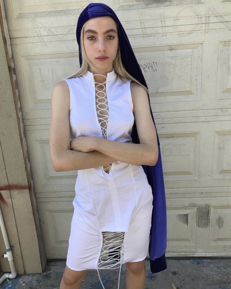 Model against a scratched white garage door wearing a white lace-up sleeveless top and lace-up shorts with a violet head drape