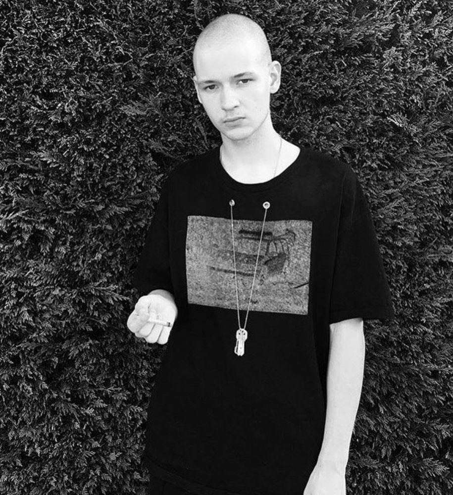 Black-and-white photograph against a dense conifer hedge: model in a black tee carrying a rectangular halftone photo print held at the shoulders by safety pins, a key pendant on a long chain