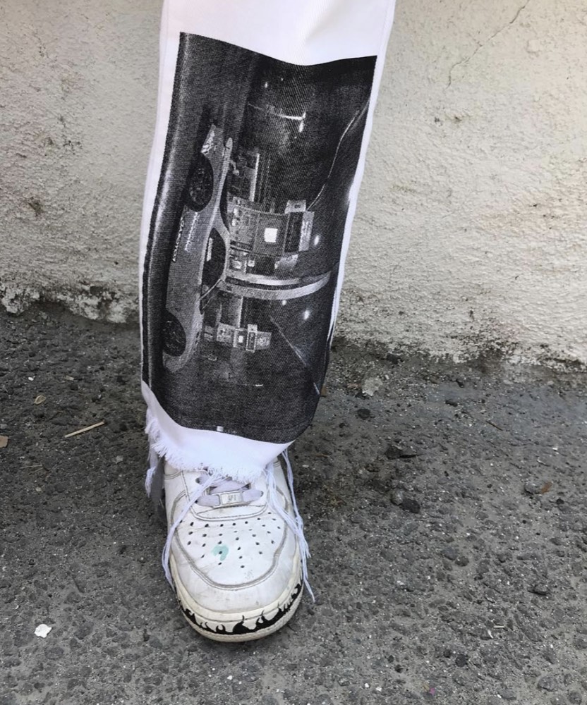 Detail photograph: white denim leg carrying a large grainy halftone photo panel, hem left raw and frayed above a worn white sneaker on cracked asphalt