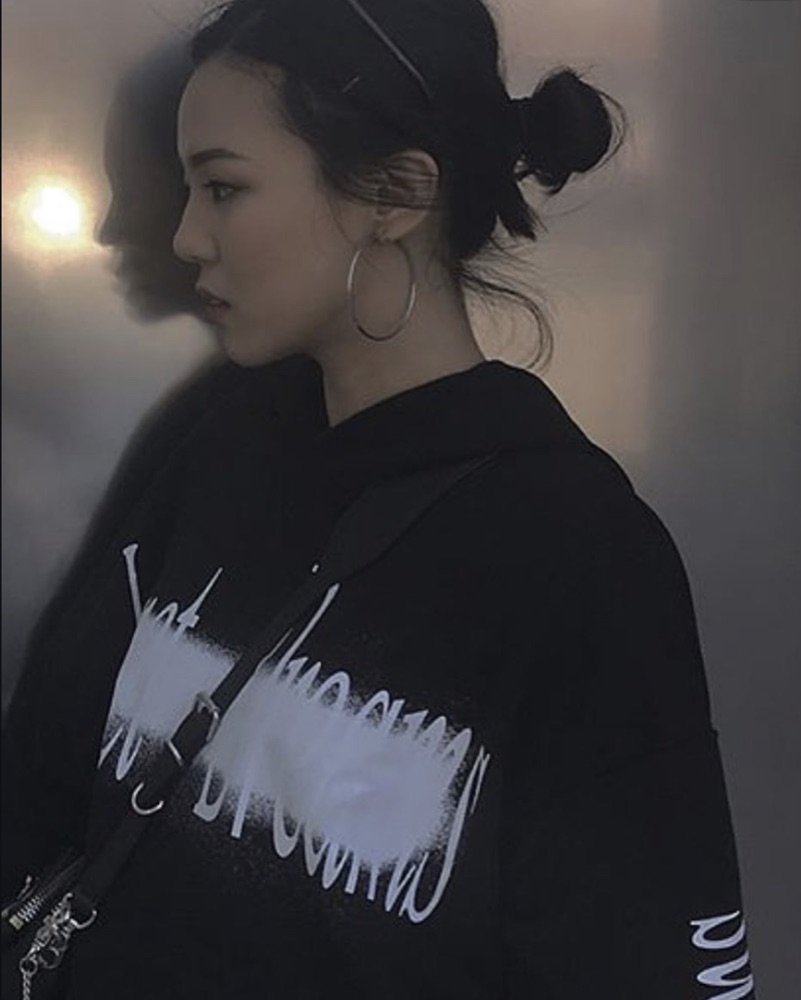 Black-and-white photograph in profile: model in a black hooded sweatshirt carrying a wide white sprayed script across the chest and sleeve, hoop earring, mist behind