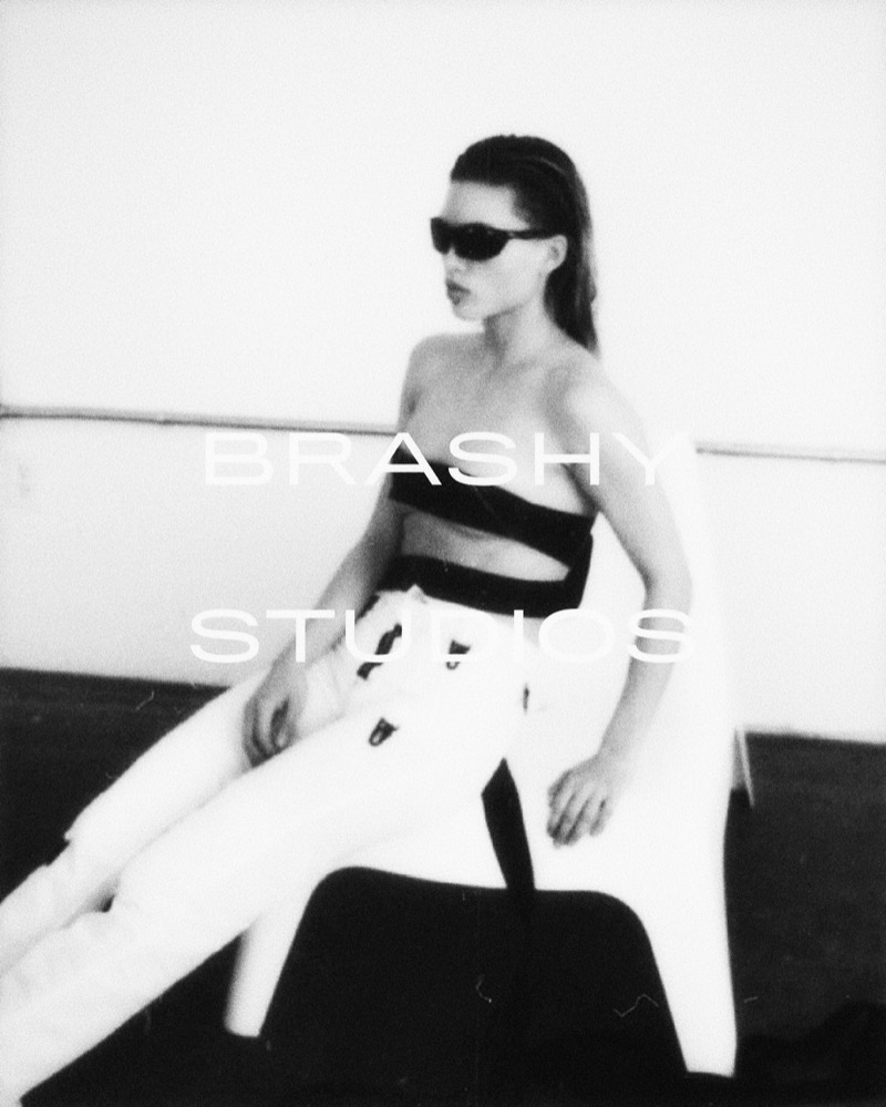 Black-and-white lookbook photograph, model seated wearing dark glasses