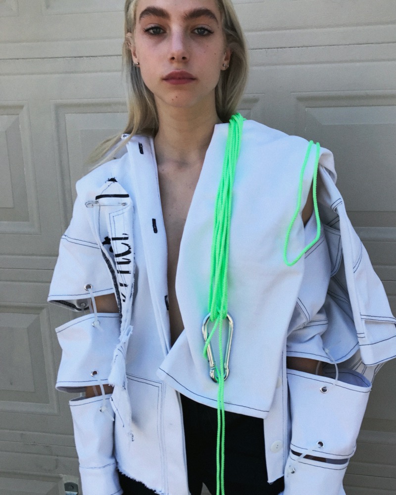 Model in a deconstructed white jacket with neon green cord and carabiner, photographed against a garage door