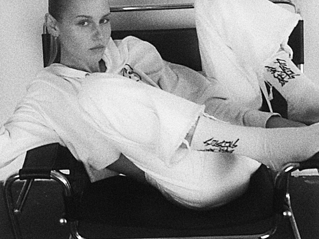 Black-and-white VHS still: model reclined on a tubular steel chair